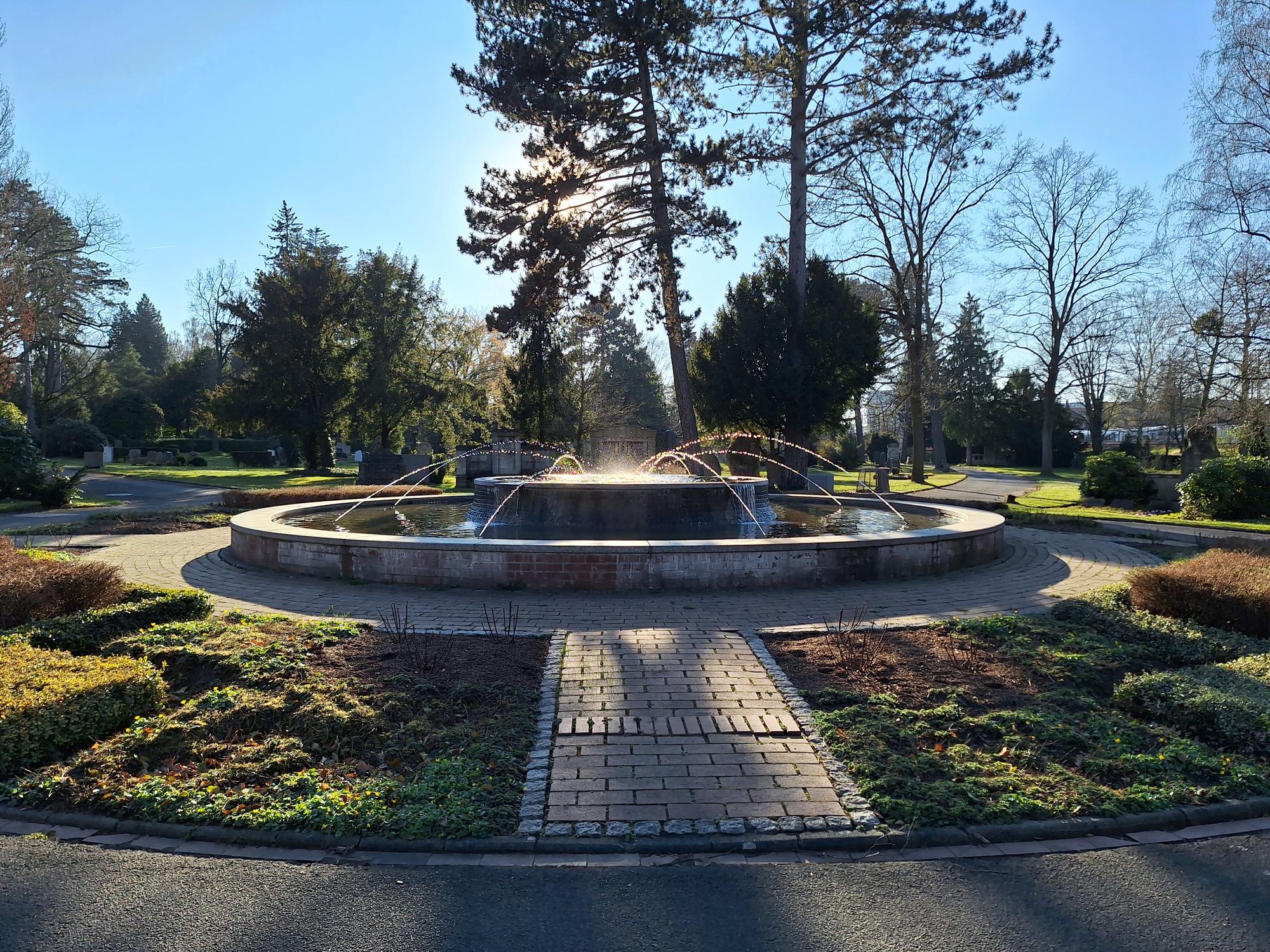 

AI generated caption: Fountain with surrounding trees and sunlight in a park setting.
