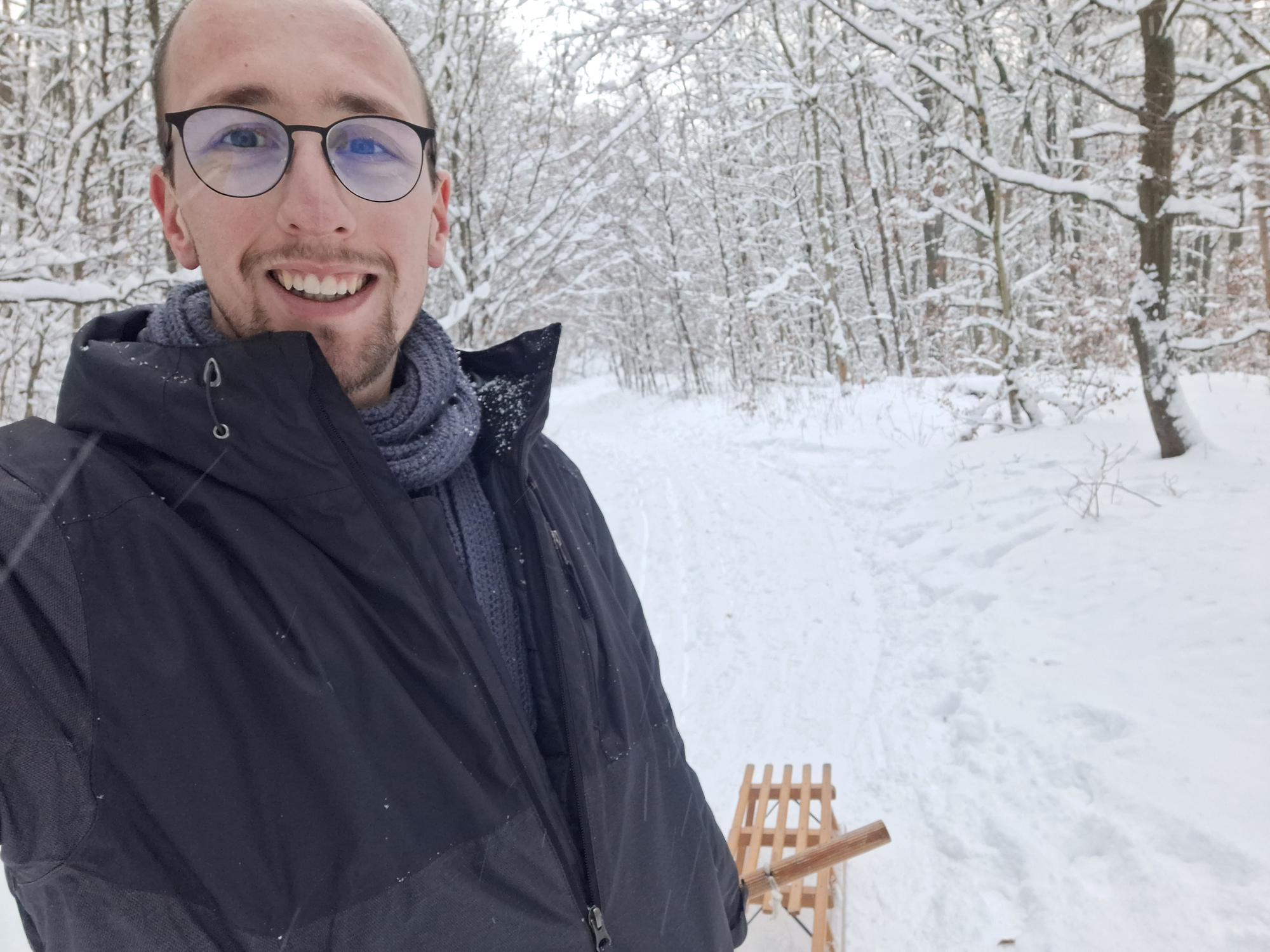 

AI generated caption: Man in glasses and winter coat with a sled in snowy forest.